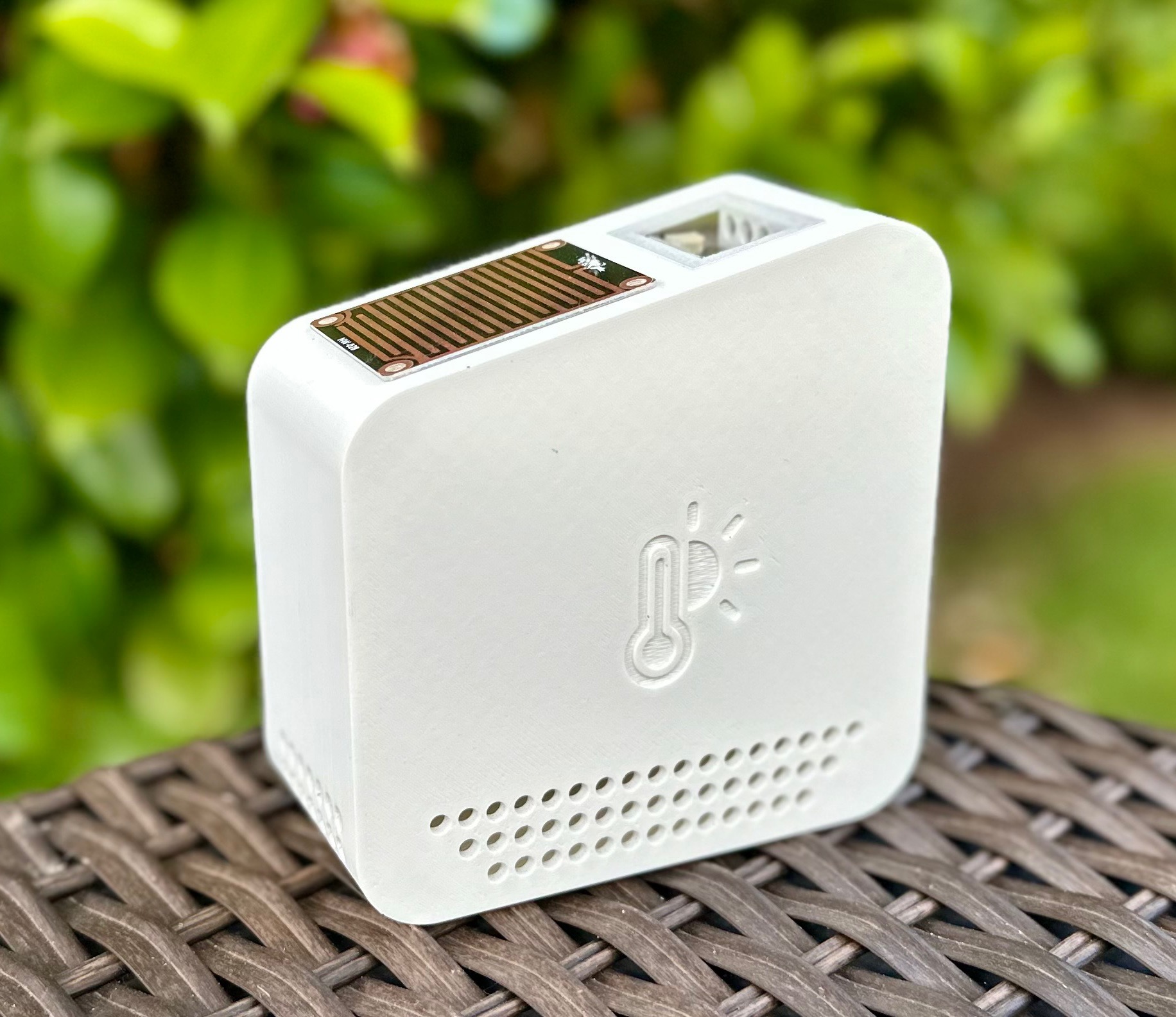The MyWeather outdoor sensor, a small white enclosure with an embossed thermometer-and-sun icon, mounted on a wooden post in a garden bed
